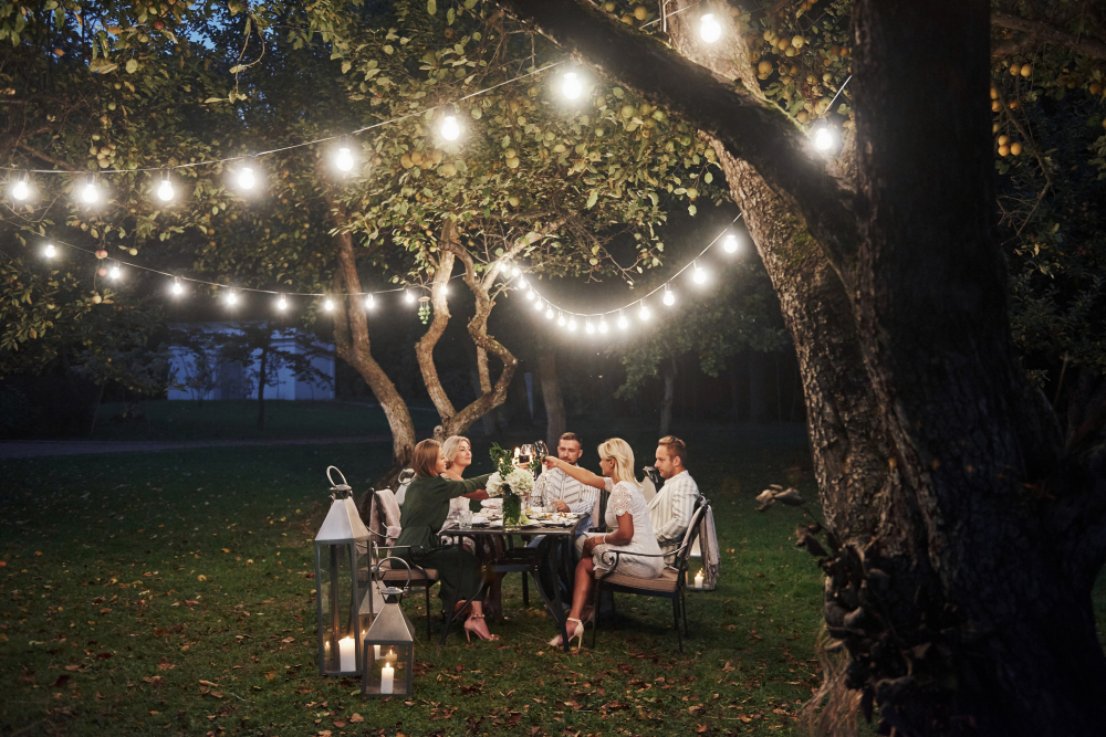 A group of friends enjoying a cozy dinner on a beautifully decorated patio under string lights in a park in Indio, CA. The table is set with food, and they are laughing and toasting together