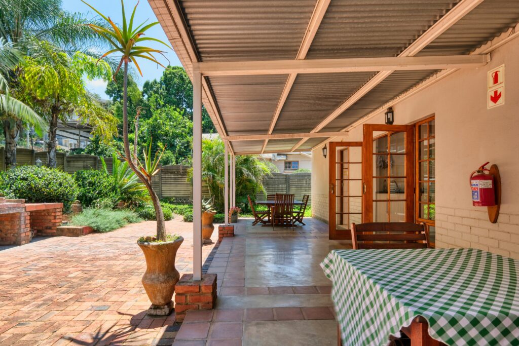 Covered outdoor patio with fixed-frame awning, brick flooring, potted plants, and wooden dining furniture, surrounded by lush greenery.