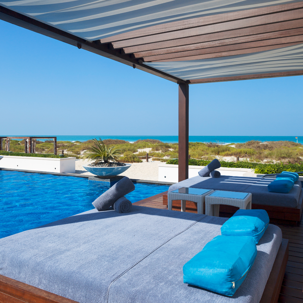 Luxurious cantilevered pergola by a serene poolside with cushioned lounge beds and a breathtaking ocean view.