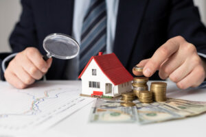 Man examining coins with a magnifying glass on a table, next to a model house labeled "CS PATIO COVER